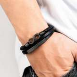 Infinity Leather Bracelet For Men - Black wrist wearing a black multi-strand leather bracelet with a silver-toned infinity symbol and a polished rectangular metal bar.